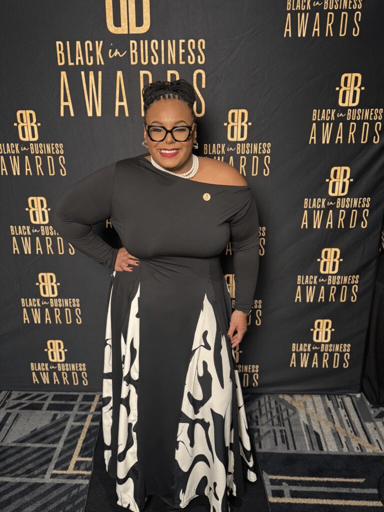 A woman in a black and white dress poses in front of a "Black in Business Awards" step-and-repeat banner on a patterned carpet.