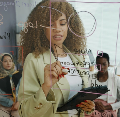 A woman writes on a glass board with diagrams and notes while holding a tablet, with two colleagues seated in the background.