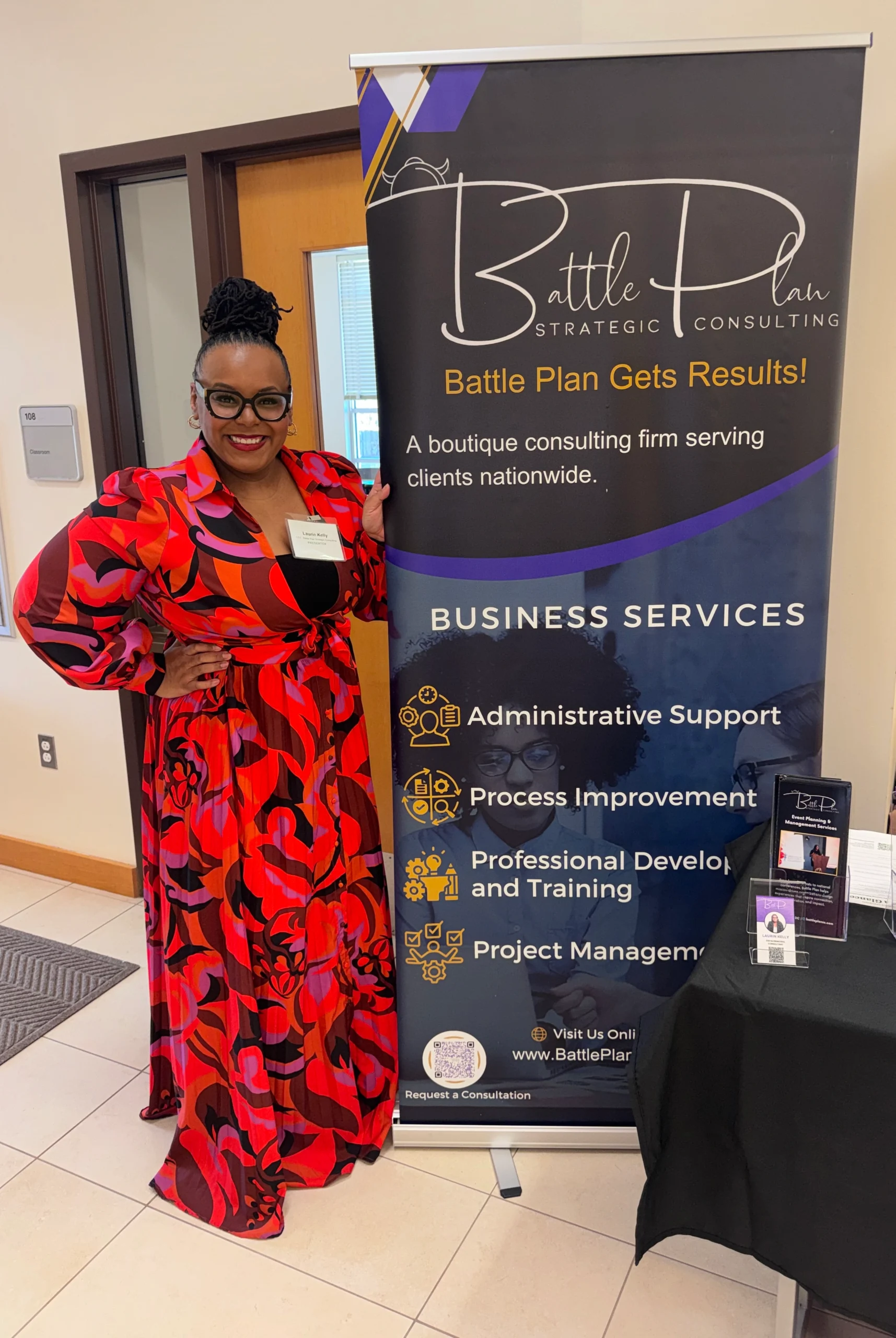 Woman in a red and black patterned dress stands next to a "Battle Plan Strategic Consulting" banner advertising business services, including administrative support and project management.