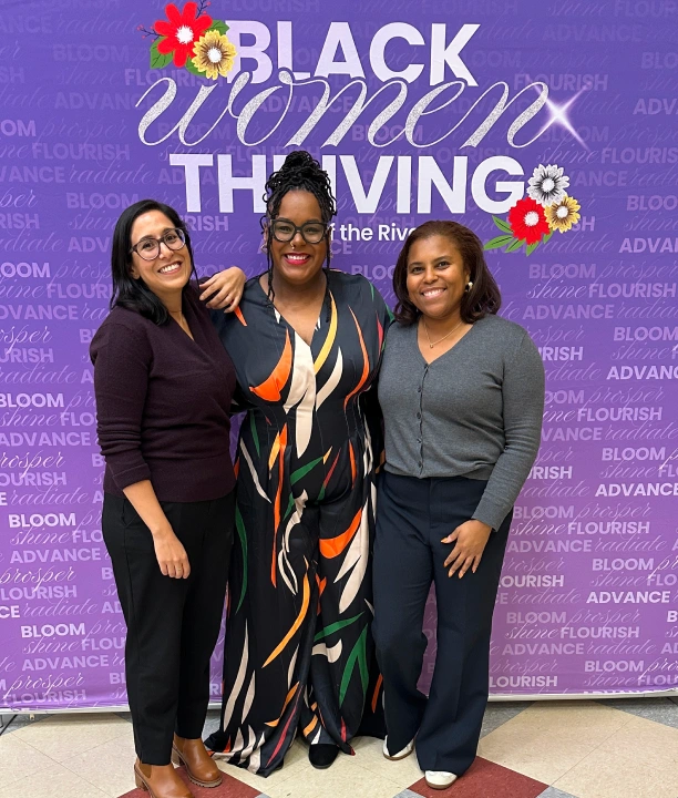 Three women stand and smile in front of a purple backdrop that reads "Black Women Thriving" decorated with floral graphics.