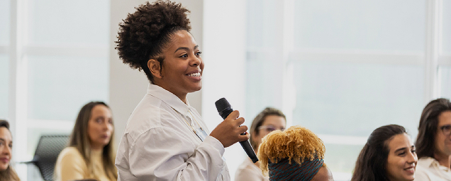 A woman stands holding a microphone and smiling while speaking to a group of seated people in a bright room.